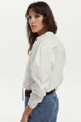 Blusa de algodón liviano color blanco, con detalles de puntilla en el frente, cuello mao con escote en V, cordón colgante y mangas largas abullonadas con puños ajustados.