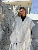 Ruana tejida en lana merino, color beige y crudo, con diseño de trenzas verticales en el frente y terminación con flecos en el ruedo.