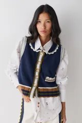 Chaleco de lana con cierre frontal de botones, cuello estilo bebé con borde tejido, y detalle de bolsillo frontal. Presenta un diseño en color azul marino con franjas horizontales en tonos marrón, verde y blanco en la parte inferior y en los bordes.