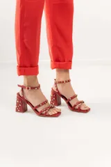 Sandalias de tacón ancho de 7 cm, color bordó, con diseño de tiras cruzadas en el empeine y pulsera al tobillo con cierre de hebilla. El modelo está decorado con tachas metálicas plateadas y doradas distribuidas por todas las tiras y el tacón.