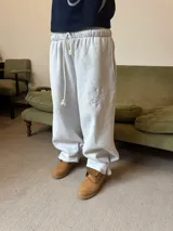 Pantalón de jogging gris melange de corte baggy, confeccionado en algodón. Presenta cintura elástica con cordón largo ajustable, bordado decorativo en la pierna izquierda y botones metálicos en la parte inferior de la botamanga para ajustar el ancho.