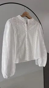 Blusa de brodery color blanco, con cuello redondo, mangas largas abullonadas con puños elásticos y cierre frontal con botones forrados. Presenta detalles de puntillas verticales en el frente.