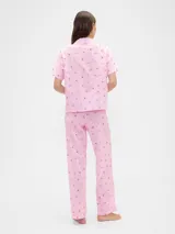 Pijama de dos piezas confeccionado en popelina, compuesto por una camisa de manga corta con cuello solapa y cierre frontal de botones, y un pantalón largo de corte recto con cintura elástica. Presenta un estampado integral de cerezas sobre fondo rosado.