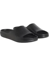 Ojotas tipo slide negras, con tira ancha que presenta el logo 'CK' en relieve en el lateral. La plantilla es contorneada con textura acanalada para mayor agarre.