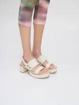 Sandalias de plataforma con taco cuadrado de 6 cm y base de 3 cm. Presentan dos tiras anchas en el empeine con detalles de doble argolla metálica en los laterales y una correa trasera con elástico para mayor ajuste.