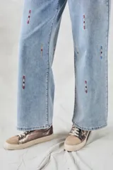 Pantalón de jean celeste de corte ancho, con bordados verticales de líneas rojas, blancas y azules.