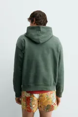 Sudadera con capucha de corte relajado, color verde oscuro, con interior perchado y bordado pequeño de una silla en el centro del pecho.