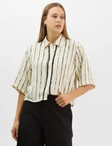 Camisa de corte holgado y silueta recta, con estampado de rayas verticales irregulares en color negro sobre fondo crema. Presenta cuello camisero clásico, mangas cortas amplias y cierre frontal con botones.