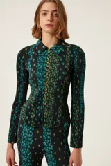 Camisa plisada negra con estampado floral en tonos verdes y azules, cuello camisero, botones dorados y mangas largas acampanadas.