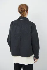 Chaqueta de paño color gris oscuro, con solapas amplias y cierre frontal mediante un alamar único. Presenta bolsillos laterales y mangas largas con puños rectos.