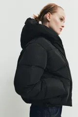 Campera puffer acolchada de color negro, con cuello envolvente, capucha integrada, manga larga, bolsillos laterales ocultos y bajo con elástico. Cierre frontal con botones a presión ocultos por solapa.