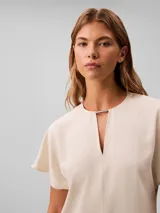 Blusa de manga corta Calvin Klein en color blanco marfil, confeccionada en tejido de crepé de chine. Presenta un diseño de corte regular con cuello en V adornado con un detalle de barra metálica horizontal.