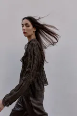 Blusa de manga larga con cuello redondo, confeccionada en tejido semitransparente con estampado paisley en tonos marrones oscuros. Presenta detalles de volantes y apliques de terciopelo en el frente y hombros, con cierre frontal de botones forrados.