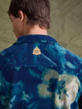 Sobrecamisa de tejido polar con estampado abstracto en tonos rojizos y azul marino. Presenta cuello clásico, cierre frontal con botones a presión, dos bolsillos frontales con solapa en el pecho y un parche decorativo.