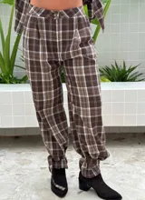 Pantalón de tiro alto con diseño a cuadros en tonos marrón y blanco. Presenta pinzas en la cintura, corte holgado y detalle de ajuste con trabilla en los tobillos.
