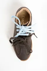 Zapatilla de cuero marrón oscuro, estilo deportivo retro, con suela de goma color caramelo. Presenta costuras decorativas en el cuerpo y el nombre "LEMON" grabado en dorado en el lateral. Se muestra con dos cordones anudados: uno celeste claro y otro marrón oscuro.