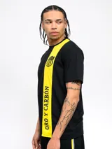 Remera amarilla con una franja vertical negra en el lado izquierdo. En la parte superior de la franja se encuentra el escudo del Club Atlético Peñarol. En la parte inferior de la franja se lee la leyenda "Oro y Carbón".