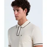 Polo color crema de algodón piqué, con cuello tipo camisa y cierre de botones. Tiene mangas cortas con ribete negro, bolsillo de parche en el pecho y logo bordado de pingüino.