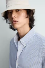 Camisa de algodón de corte relajado, con cuello solapa abotonado (button-down) y manga larga terminada en puño con botón. Presenta un patrón de rayas verticales finas en tonos azul claro y blanco.