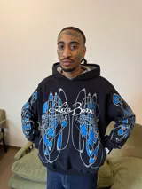 Hoodie oversize de color negro con estampado artístico en pecho y mangas en tonos azul y gris. Presenta capucha forrada con tejido a cuadros, costuras reforzadas y grifa de la marca en la manga.