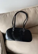 Cartera de hombro tipo baguette en color negro con acabado brillante efecto cuero arrugado. Presenta una estructura rectangular con bordes redondeados, cierre superior y doble asa fija.