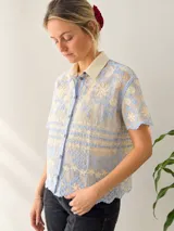 Camisa de manga corta confeccionada en tejido transparente con bordados florales en tonos celeste y crema, cuello clásico, cierre frontal con botones y terminaciones festoneadas.