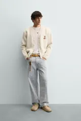 Cárdigan de punto color beige con cuello en pico, manga larga y cierre frontal de botones. Presenta un pequeño bordado a contraste en el pecho y acabados en rib en puños y bajo.
