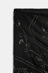 Pantalón bombacho negro semitransparente de tiro medio y cintura elástica. Tiene un pareo superpuesto ajustable con lazada lateral, decorado con abalorios, lentejuelas y flecos plateados. El bajo es elástico.