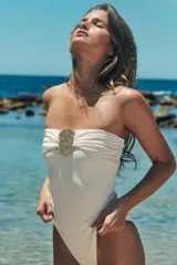 Bañador enterizo tipo bandeau color crema, con escote recto drapeado y detalle de pieza metálica circular dorada en el centro del pecho. Cuenta con forro interior.