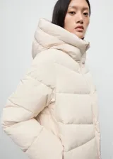 Campera acolchada de corte recto y diseño puffer, color beige, con capucha, cuello alto y cierre frontal de cremallera doble. Cuenta con mangas largas con puños elásticos y dos bolsillos laterales con cierre.
