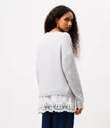 Suéter de punto color gris con escote en V y mangas largas. Presenta un diseño alargado con un detalle de encaje blanco en el ruedo inferior.