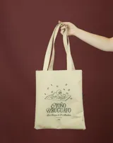 Tote bag de color beige con estampado gráfico en color verde que ilustra un perro con lentes sobre una manta, acompañado por la frase 'Otoño Uruguayo'. Cuenta con doble asa larga para llevar al hombro.
