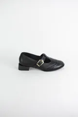 Zapato tipo guillermina estilo oxford, confeccionado en cuero negro con detalles de perforaciones decorativas en la puntera y cierre mediante tira con hebilla metálica dorada. Presenta un taco bajo cuadrado.