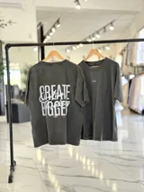 Remera oversize de color gris con efecto gastado (acid wash). Presenta un diseño de tipografía en la parte frontal y un gran estampado en la espalda con la inscripción "Create Bigger". Cuenta con mangas cortas y un calce holgado y cómodo.