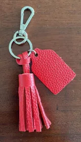 Llavero de cuero rojo con borla y etiqueta rectangular para grabado. Incluye gancho mosquetón y argolla metálica plateada.