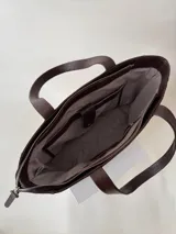 Cartera tipo tote de cuero graneado negro, con diseño clásico y textura uniforme. Cuenta con doble asa de hombro, bolsillo frontal con cierre magnético, bolsillo trasero con cierre metálico y compartimento principal con cierre. El interior está forrado y dispone de múltiples bolsillos organizadores.