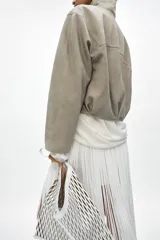 Chaqueta corta de ante con cuello alto, manga larga y cierre frontal con cremallera y botones a presión. Presenta bolsillos frontales con solapa y bajo abullonado con pliegues.