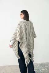 Ruana de lana en color crudo con diseño de rayas verticales en tonos tierra y detalles sutiles de hilo brillante. Presenta terminaciones con flecos en el ruedo y una etiqueta de cuero en la parte inferior.