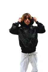 Hoodie con cierre frontal, color negro, con estampado gráfico de hojas de acanto en color blanco que recorre el frente y las mangas. Cuenta con bolsillos laterales y cierre metálico.