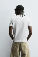 Camiseta de corte relajado, color gris claro, confeccionada en tejido con estructura waffle. Presenta cuello redondo y manga corta, con costuras decorativas en contraste color dorado que recorren los laterales y los hombros.