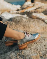 Zapato acordonado estilo oxford de cuero vacuno en color plateado metalizado, con diseño brogue, suela de goma y taco de madera.