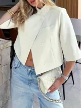 Chaqueta cropped de diseño estructurado en color crema, con cuello alto, cierre frontal cruzado con botones y mangas 3/4. Presenta un bolsillo decorativo en el frente y un corte asimétrico.