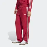 Pantalón deportivo Adidas Firebird Drop Needle de color rojo con rayas verticales finas, bolsillos laterales con cierre y las icónicas tres tiras blancas a lo largo de los laterales. Incluye el logo del trifolio bordado en la pierna.