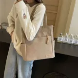 Cartera tipo shopper color beige con bolsillos laterales y un bolsillo frontal con tirador de cordón trenzado. Cuenta con asas superiores de lona.