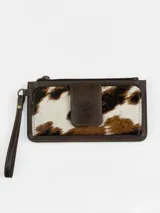 Billetera porta celular confeccionada en cuero marrón con detalle de pelo vacuno con estampado animal print tipo vaca en tonos blancos, marrones y negros. Cuenta con correa de muñeca y cierre superior.