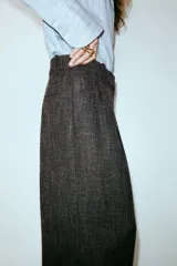 Pantalón sastrero de tiro alto, color marrón oscuro con textura tweed o espigada. Presenta un corte amplio y recto, con pinzas marcadas en la parte delantera.