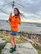 Chaqueta impermeable naranja con cuello alto y detalles de volados en color verde oliva. Presenta cierres y bolsillos frontales, puños ajustados y cordón regulable en la cintura.