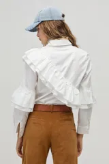 Camisa blanca de popelina de algodón con cuello clásico y botones de nácar. Presenta volados dobles decorativos que recorren el frente y la espalda desde el hombro hasta la cintura, además de volados en las mangas a la altura del codo.