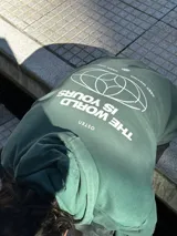 Canguro verde con capucha y cordones ajustables. Presenta un bolsillo tipo canguro y dos estampados blancos: un logo con forma de globo terráqueo en el lado izquierdo del pecho y una flor en el lado derecho.