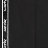 Pantalón deportivo Supreme de color negro, confeccionado en poliéster. Presenta una banda lateral con el logo de la marca repetido y el logo de Supreme bordado en la parte superior delantera. Cuenta con cintura elástica y puños ajustados en los tobillos.